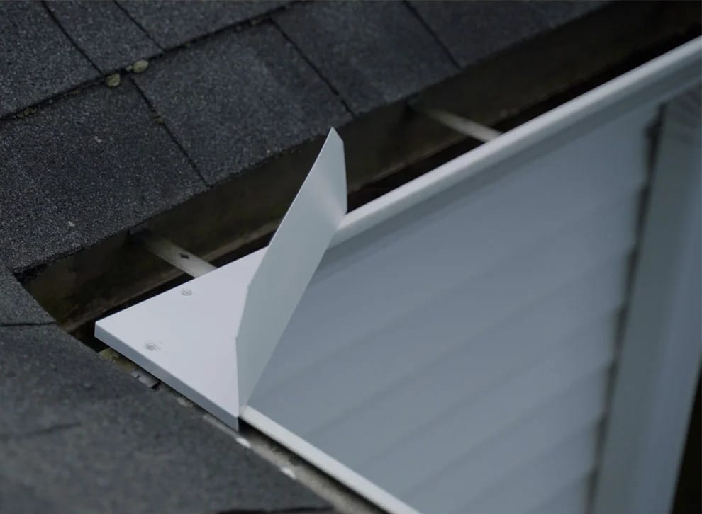 Close-up of a metal flashing installed along the edge of a shingled roof, showing the junction between the roof and a vertical siding wall.