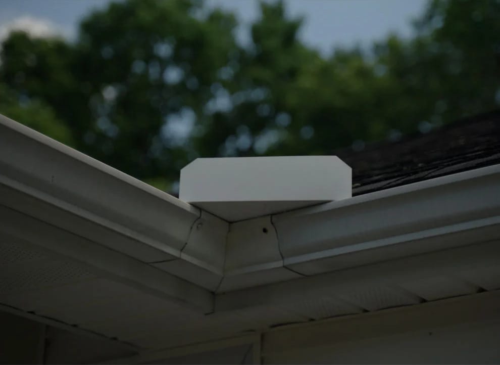 A white electronic device is installed at the corner of a house roof near the gutter, with trees visible in the background.